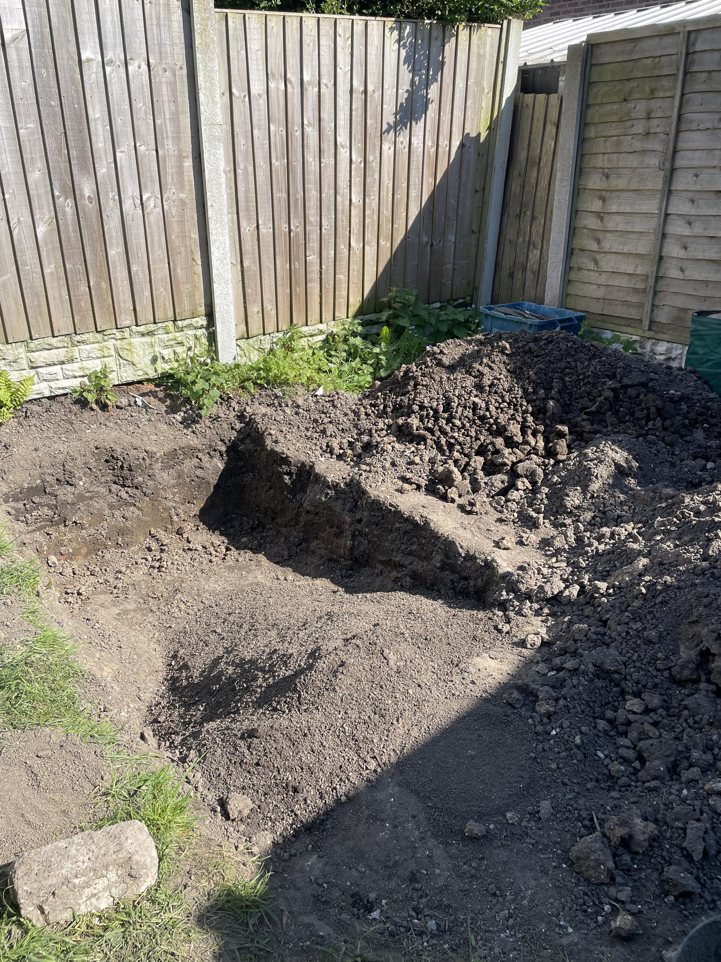 A garden surrounded by fence panels with a large deep hole dug into the ground surrounded by piles of soil