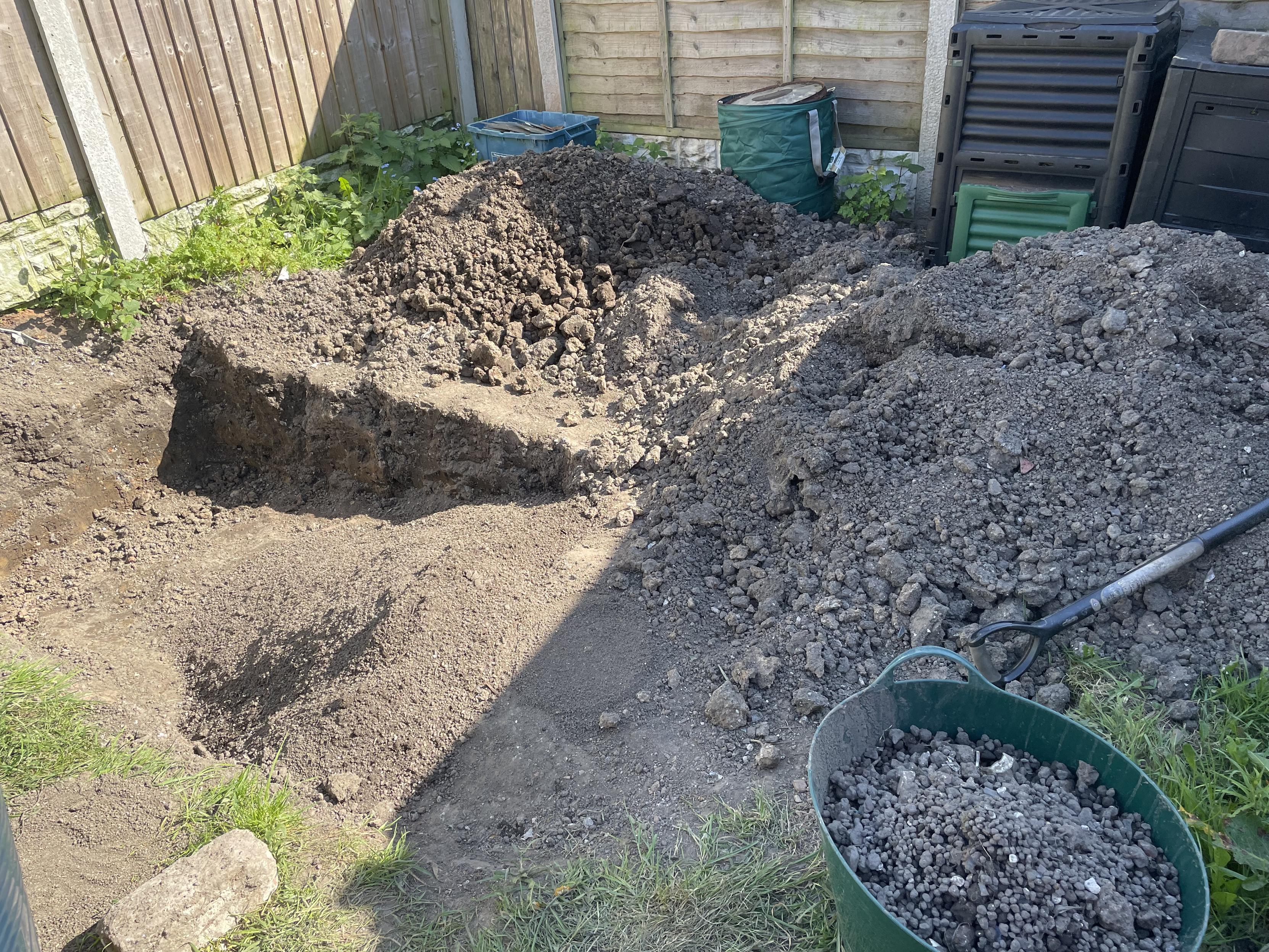 A garden surrounded by fence panels with a large hole dug into the ground surrounded by piles of soil and a bucket with rubble in